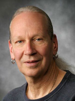 Jim Struve, a Light-skinned man with hair pulled back and double earings in front of a grey wall.