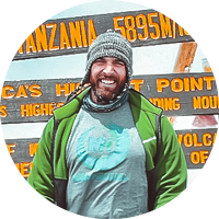 A smiling, bearded man in front of a sign showing him at the peak of Mount Kilimanjaro.